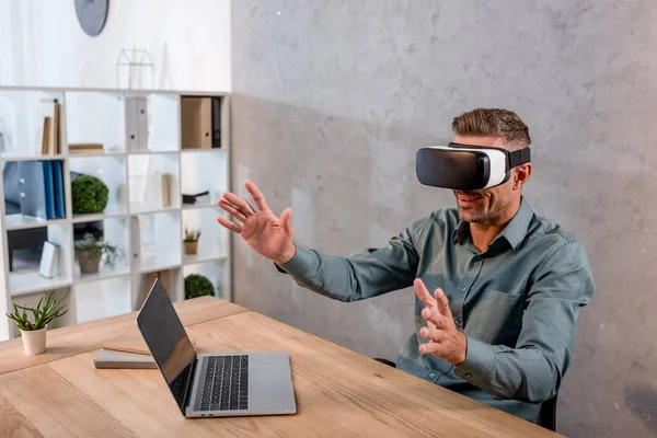 Homme d'affaires portant un casque de réalité virtuelle assis au bureau près d'un ordinateur portable — Photo de stock