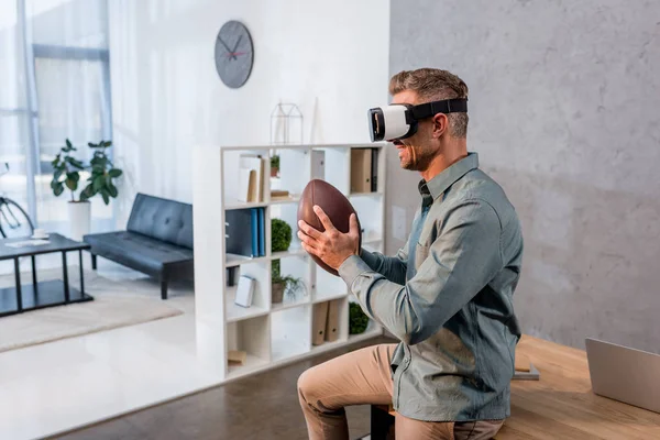 Alegre hombre de negocios con auriculares de realidad virtual mientras sostiene el fútbol americano - foto de stock