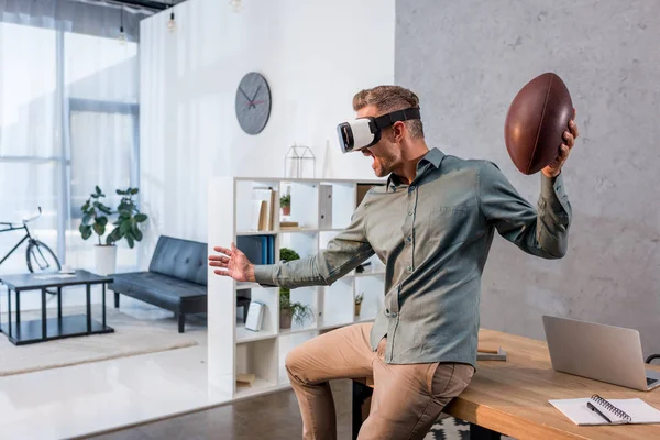 Homem de negócios feliz vestindo fone de ouvido realidade virtual e segurando futebol americano — Fotografia de Stock