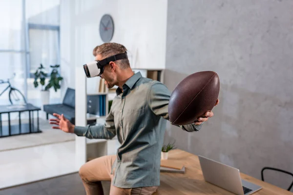 Empresário vestindo realidade virtual headset e segurando futebol americano em offcie — Fotografia de Stock