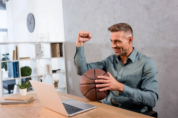 Fröhlicher Geschäftsmann, der Basketball hält, während er im Büro auf dem Laptop die Meisterschaft verfolgt — Stockfoto