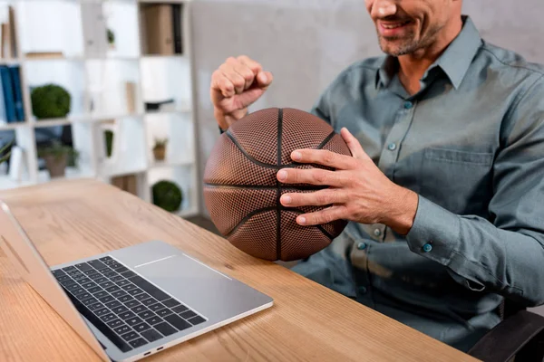 Vue recadrée de l'homme d'affaires joyeux tenant basket tout en regardant le championnat sur ordinateur portable au bureau — Photo de stock