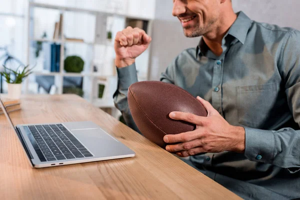 Vista cortada de empresário alegre assistindo campeonato no laptop e segurando futebol americano no escritório moderno — Fotografia de Stock