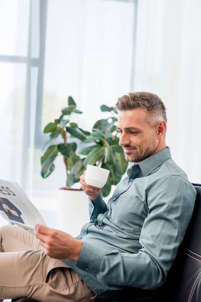 Happy businessman sitting on sofa and holding cup while reading newspaper — Stock Photo