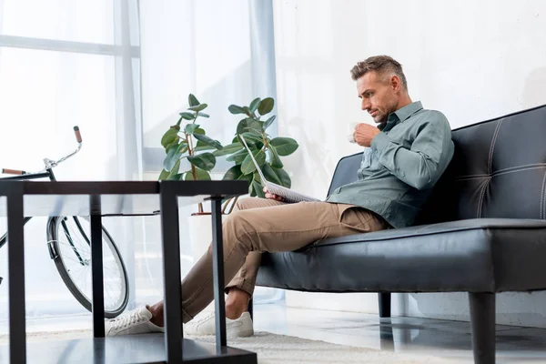 Businessman sitting on sofa and holding cup with drink while reading newspaper — Stock Photo