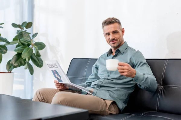 Fröhlicher Geschäftsmann auf Sofa sitzend, während er Wirtschaftszeitung und Tasse mit Getränk in der Hand hält — Stockfoto