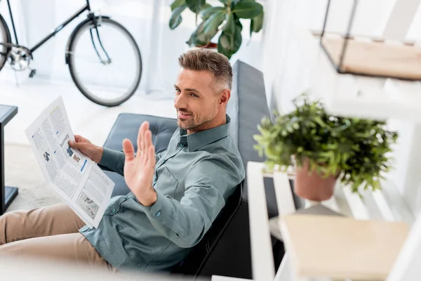 Selective focus of cheerful businessman holding newspaper and waving hand while sitting on sofa — Stock Photo