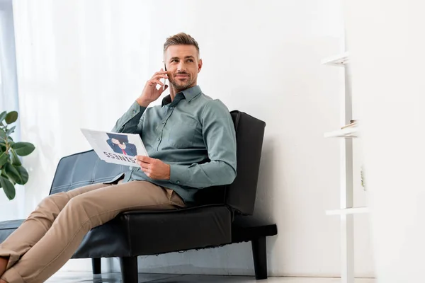 Cheerful businessman holding business newspaper and talking on smartphone while sitting on sofa — Stock Photo