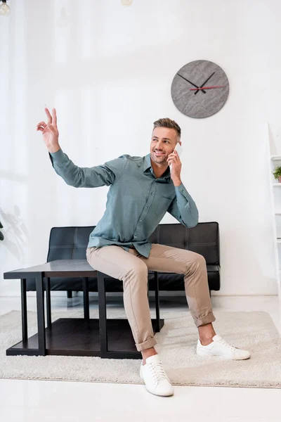 Cheerful businessman sitting on coffee table and showing peace sing while talking on smartphone — Stock Photo