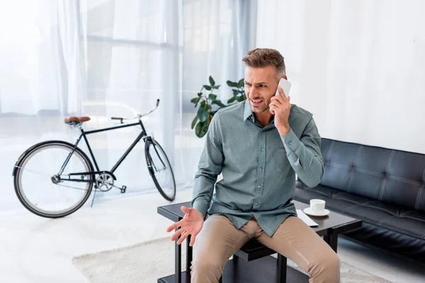 Emotional businessman talking on smartphone while sitting on coffee table in office — Stock Photo