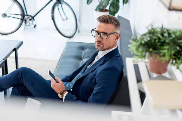 Selective focus of businessman in glasses sitting on sofa and holding smartphone while looking at camera — Stock Photo