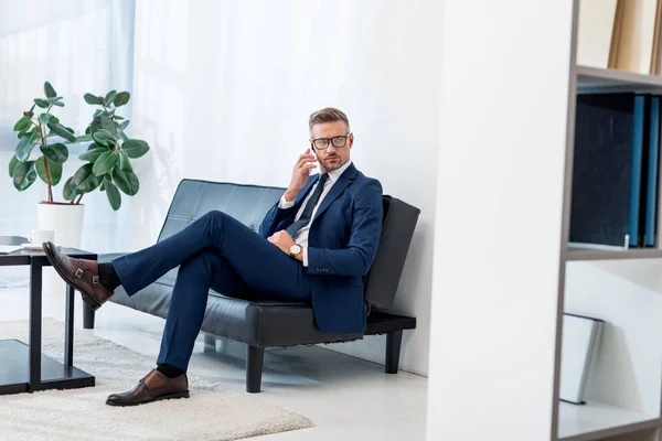 Homem de negócios bonito em óculos sentado no sofá e falando no smartphone — Fotografia de Stock