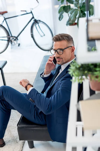 Cheerful businessman in glasses sitting on sofa and talking on smartphone while looking at camera — Stock Photo