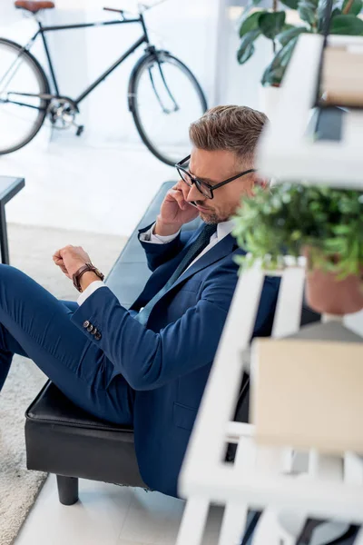 Homme d'affaires dans des lunettes assis sur le canapé et parler sur smartphone tout en regardant regarder — Photo de stock