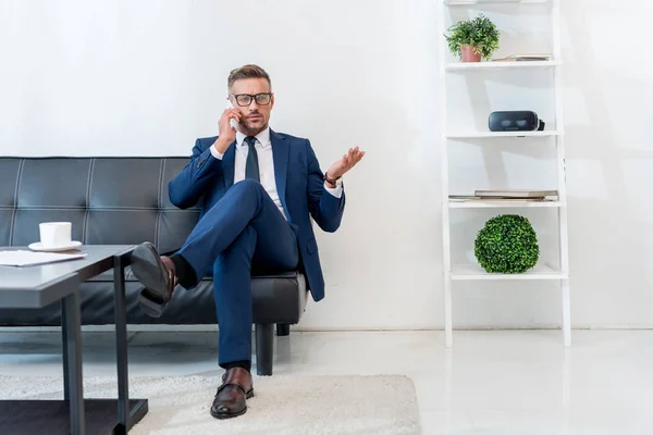 Geschäftsmann mit Brille sitzt auf Sofa und spricht auf Smartphone neben Couchtisch — Stockfoto