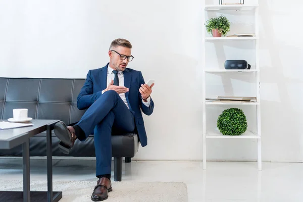 Hombre de negocios usando el teléfono inteligente y el gesto mientras está sentado en el sofá - foto de stock