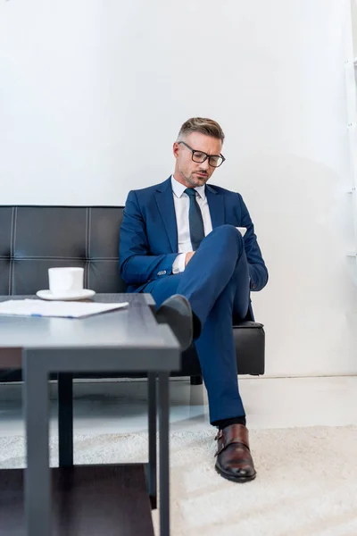 Selektiver Fokus eines gutaussehenden Geschäftsmannes in Brille, der auf einem Sofa mit überkreuzten Beinen am Couchtisch sitzt — Stockfoto