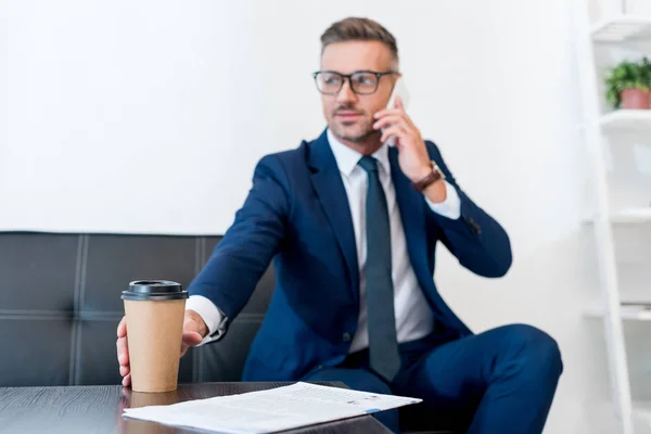 Enfoque selectivo de hombre de negocios en gafas hablando en el teléfono inteligente y sosteniendo la taza de papel - foto de stock