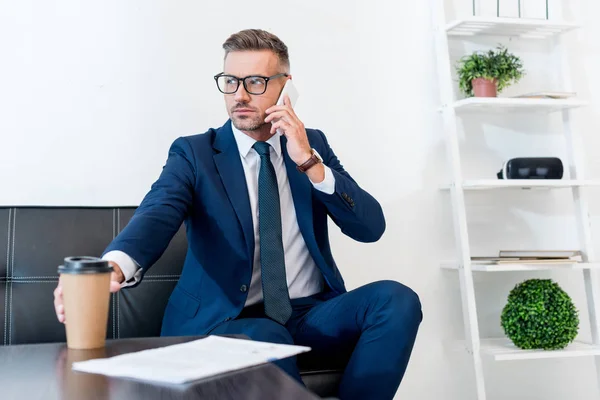 Selektiver Fokus eines gutaussehenden Geschäftsmannes mit Brille, der mit Smartphone spricht und Pappbecher hält — Stockfoto