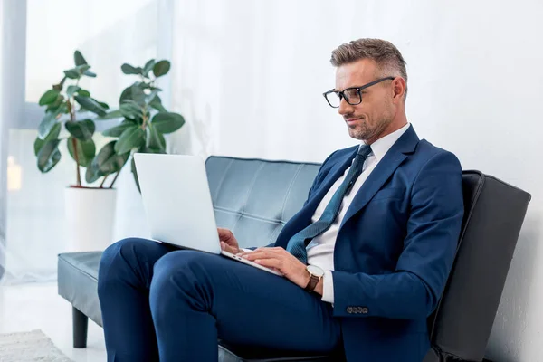 Hombre de negocios alegre en traje usando el ordenador portátil mientras está sentado en el sofá - foto de stock