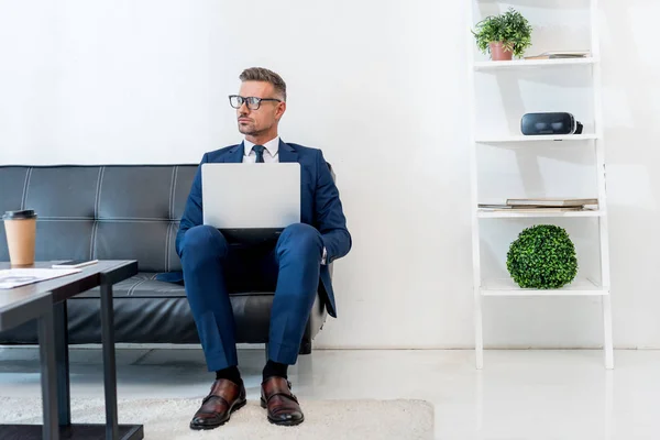 Schöner Geschäftsmann im Anzug sitzt mit Laptop auf Sofa — Stockfoto