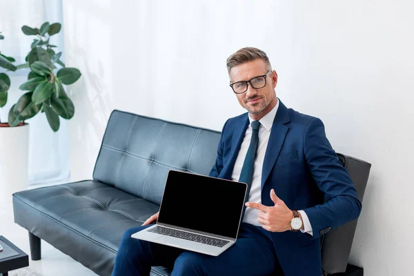 Alegre hombre de negocios en traje sentado en el sofá y apuntando con el dedo a la computadora portátil con pantalla en blanco - foto de stock