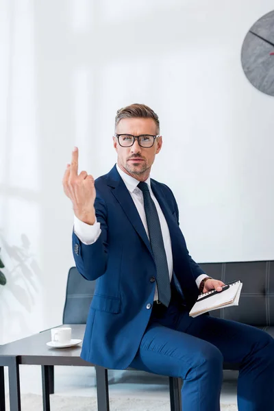 Hombre de negocios guapo en gafas que muestran el dedo medio mientras sostiene el cuaderno y la pluma - foto de stock