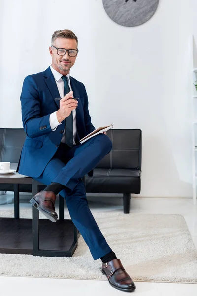 Alegre hombre de negocios en gafas que sostiene el cuaderno y la pluma mientras está sentado en la mesa de café con las piernas cruzadas - foto de stock