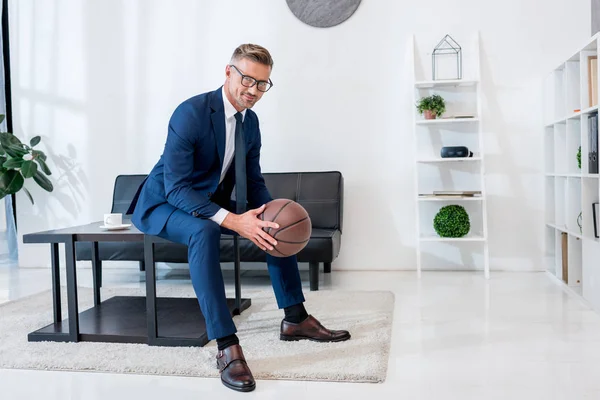 Cheerful businessman in suit holding basketball in hands while sitting on coffee table — Stock Photo