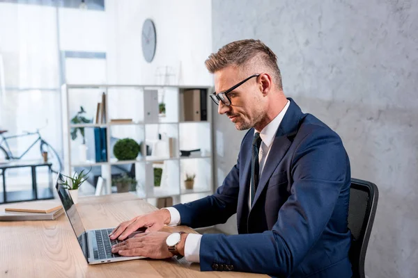 Bell'uomo d'affari in occhiali e vestito usando il computer portatile in ufficio — Foto stock