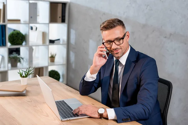 Glücklicher Geschäftsmann mit Brille, der im Büro auf dem Smartphone neben dem Laptop spricht — Stockfoto