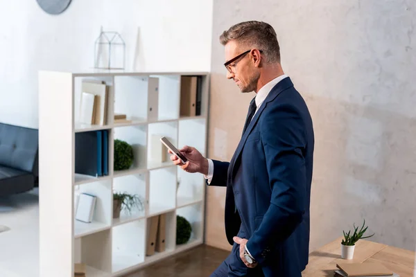 Schöner Geschäftsmann im Anzug, der mit der Hand in der Tasche steht und das Smartphone benutzt — Stockfoto