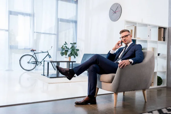 Gutaussehender Geschäftsmann mit Brille sitzt im Sessel und spricht auf dem Smartphone — Stockfoto