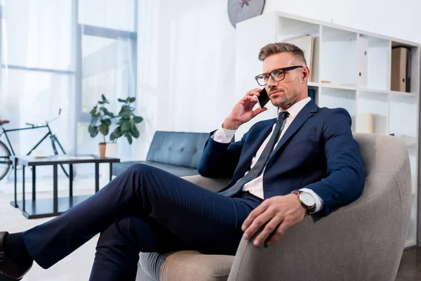Hombre de negocios en gafas sentado en sillón y hablando en el teléfono inteligente - foto de stock