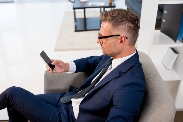 Homme d'affaires en lunettes et costume assis dans un fauteuil et regardant smartphone avec écran blanc — Photo de stock