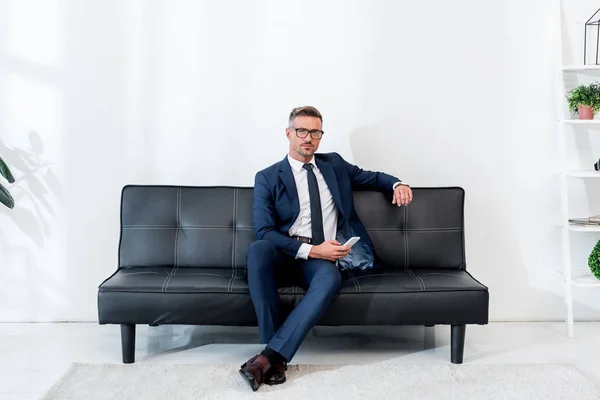 Bell'uomo d'affari in giacca e cravatta seduto sul divano e con lo smartphone — Foto stock