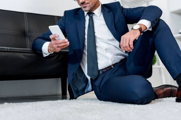 Vista recortada de hombre de negocios alegre en traje sentado en la alfombra cerca del sofá y la celebración de teléfono inteligente - foto de stock