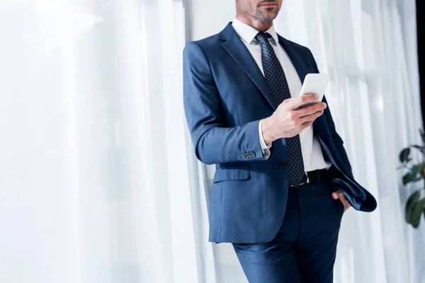 Abgeschnittene Ansicht eines Geschäftsmannes im Anzug, der mit der Hand in der Tasche steht, während er sein Smartphone benutzt — Stockfoto