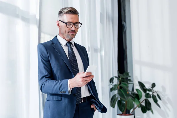 Hombre de negocios alegre en traje y gafas de pie con la mano en el bolsillo y el uso de teléfono inteligente - foto de stock