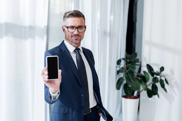Homem de negócios bonito em terno e óculos segurando smartphone com tela em branco — Fotografia de Stock