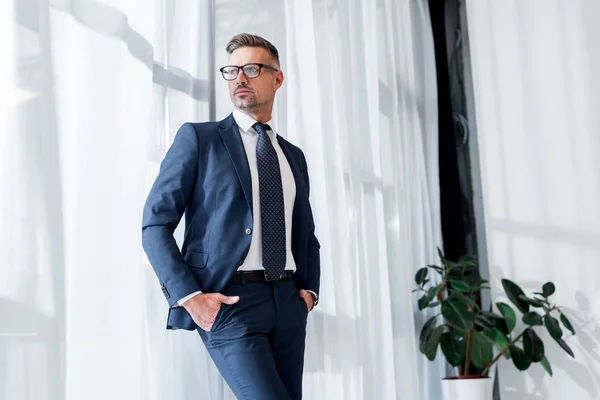 Empresario seguro de sí mismo en traje y gafas de pie con las manos en los bolsillos — Stock Photo