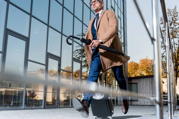 Homem de negócios bonito em óculos e casaco bege correndo com mala e guarda-chuva na rua — Fotografia de Stock