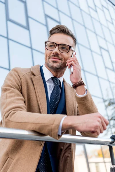 Cheerful businessman standing in beige coat and talking on smartphone — Stock Photo