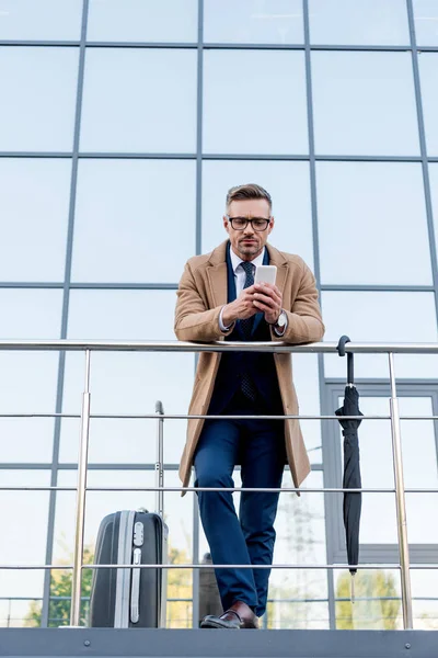 Guapo hombre de negocios con abrigo beige usando un teléfono inteligente mientras está de pie cerca de la maleta y el paraguas - foto de stock