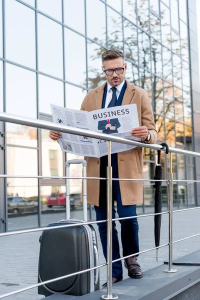 Bel homme d'affaires en manteau lecture journal d'affaires tout en se tenant près de la valise et parapluie — Photo de stock