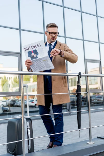 Geschäftsmann im Mantel schaut auf die Uhr, während er die Wirtschaftszeitung in der Hand hält und neben Koffer und Regenschirm steht — Stockfoto