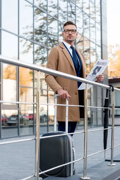 Businessman standing in beige coat and holding business newspaper and luggage — Stock Photo