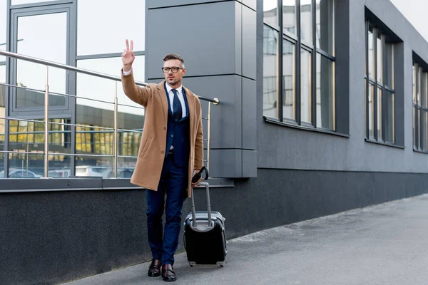 Bell'uomo d'affari in cappotto gesticolare mentre in piedi con valigia e ombrello in strada — Foto stock