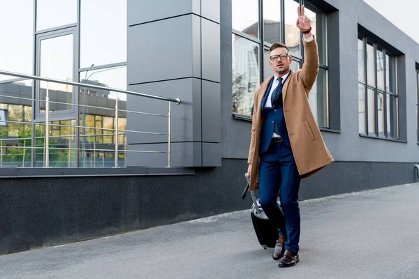 Geschäftsmann in beigem Mantel zeigt Friedenszeichen, während er mit Koffer und Regenschirm auf der Straße läuft — Stockfoto