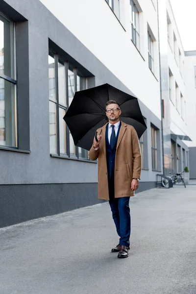 Hombre de negocios guapo en gafas caminando en abrigo beige con paraguas - foto de stock